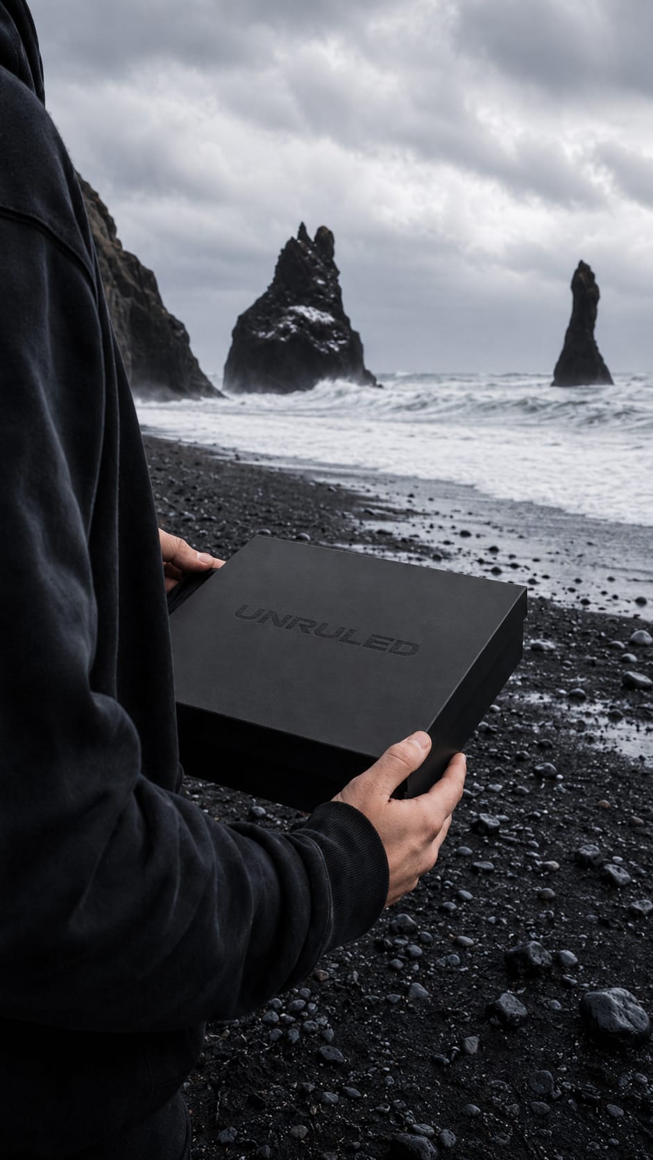 Matte black box, UNRULED debossed black on black, held on the black sand beach